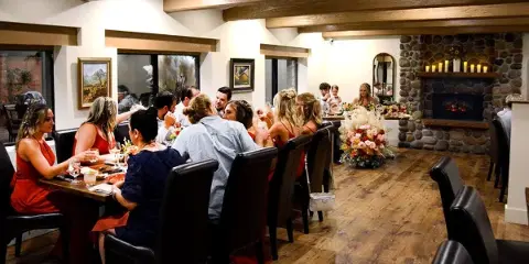 Interior view of a dining room with wood beams and a stone fireplace, showing guests seated at a long table during an event.