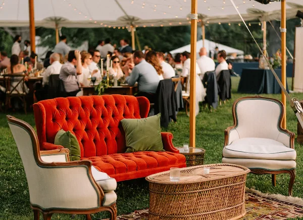 A red velvet sofa and white armchairs arranged under a white event tent with string lights at an outdoor wedding reception.