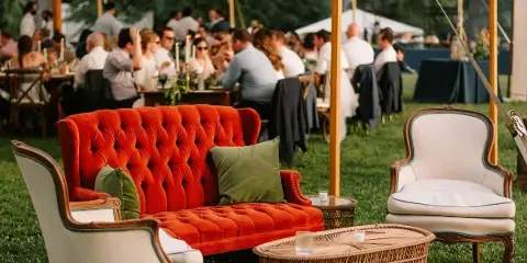 A red velvet sofa and white armchairs arranged under a white event tent with string lights at an outdoor wedding reception.
