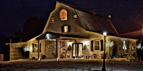 A nighttime exterior view of a large, rustic wooden lodge with a gambrel roof, illuminated windows, and a covered porch.