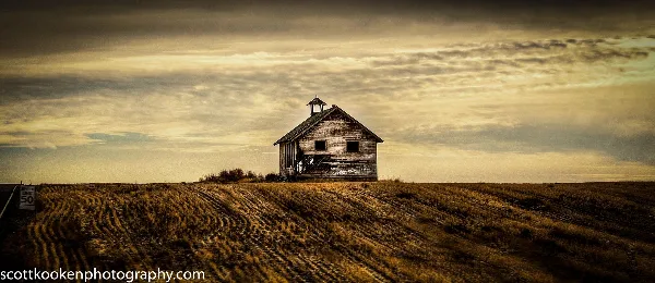 A wide landscape photograph of a weathered wooden building with a cupola situated on a grassy hill under a dramatic, cloudy sky.