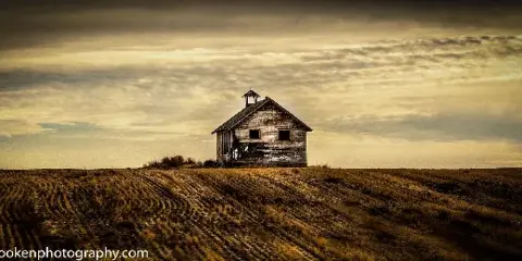 A wide landscape photograph of a weathered wooden building with a cupola situated on a grassy hill under a dramatic, cloudy sky.