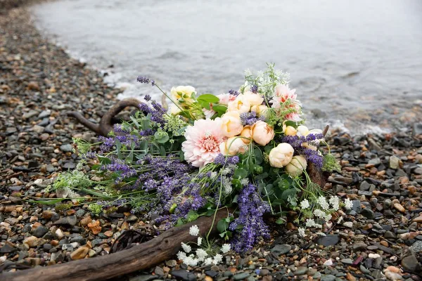A loose floral arrangement with pink peonies and purple lavender resting on a rocky beach near the water.