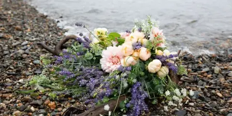 A loose floral arrangement with pink peonies and purple lavender resting on a rocky beach near the water.