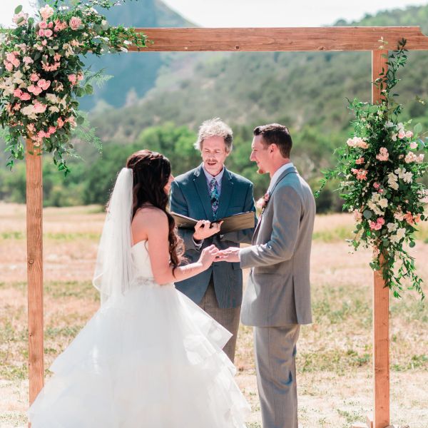 A wedding officiant presiding over an outdoor ceremony for a couple under a wooden arch decorated with flowers.