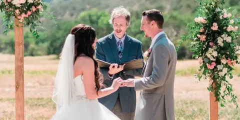 A wedding officiant presiding over an outdoor ceremony for a couple under a wooden arch decorated with flowers.
