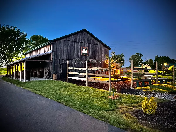 Exterior view of a rustic wooden barn wedding venue with a covered porch, string lights, and a wooden fence at dusk.