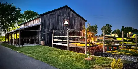 Exterior view of a rustic wooden barn wedding venue with a covered porch, string lights, and a wooden fence at dusk.