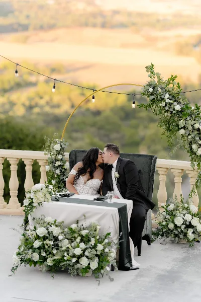 A bride and groom sharing a kiss at a wedding reception table decorated with white flowers and greenery, set against a scenic outdoor backdrop.