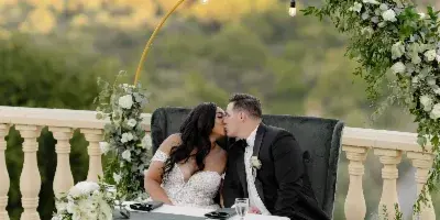 A bride and groom sharing a kiss at a wedding reception table decorated with white flowers and greenery, set against a scenic outdoor backdrop.