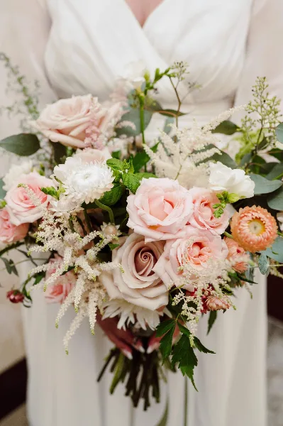 A close-up of a bride in a white dress holding a lush wedding bouquet with pink and white roses, white chrysanthemums, and greenery.