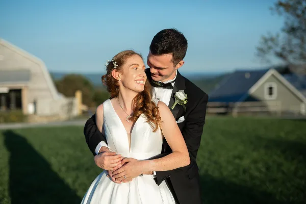 A bride and groom embracing outdoors in a field with a barn in the background.