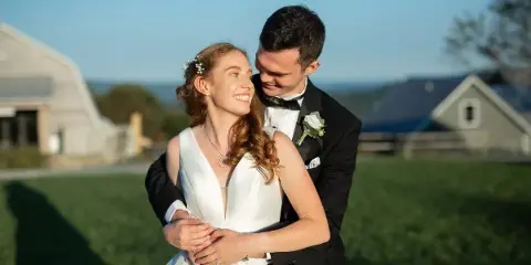 A bride and groom embracing outdoors in a field with a barn in the background.