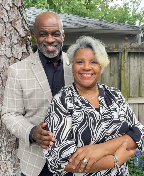 A professional portrait of a smiling man and woman standing together outdoors in front of a tree and a house.