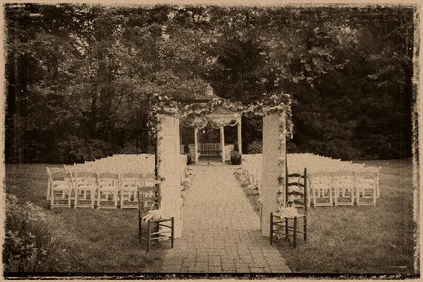 Outdoor wedding ceremony setup featuring a floral arch, rows of white chairs, and a gazebo in a garden setting.