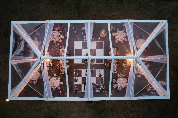 Aerial view of a clear-frame event tent with string lighting, round dining tables, and a checkered dance floor.