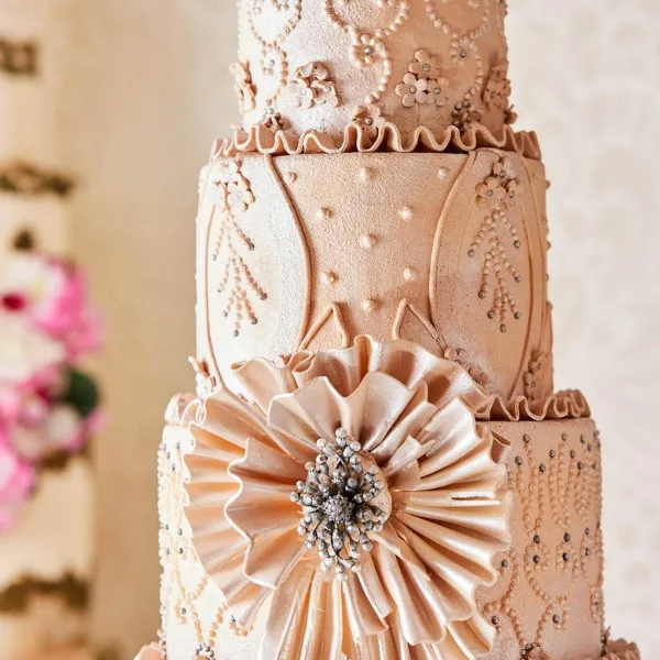 A multi-tiered wedding cake with intricate beige fondant decorations, floral patterns, and a large central flower.