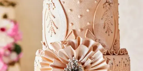 A multi-tiered wedding cake with intricate beige fondant decorations, floral patterns, and a large central flower.
