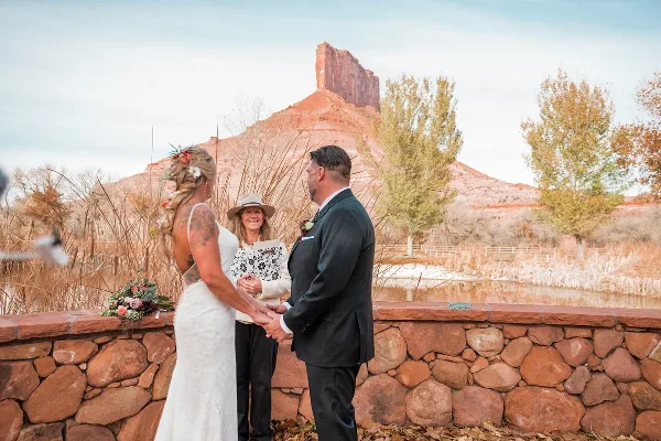 A wedding officiant stands between a bride and groom holding their hands during an outdoor ceremony.