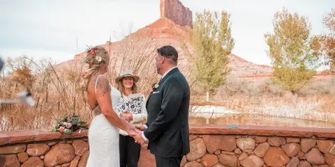 A wedding officiant stands between a bride and groom holding their hands during an outdoor ceremony.