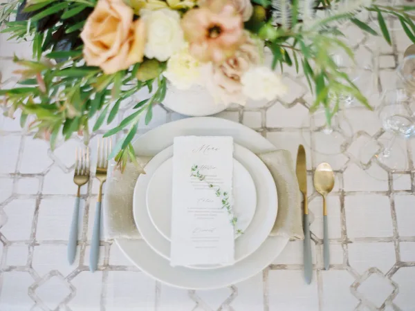 A close-up of an elegant wedding table setting featuring white plates, a custom menu card, gold and blue flatware, and a floral centerpiece.