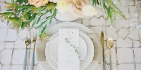 A close-up of an elegant wedding table setting featuring white plates, a custom menu card, gold and blue flatware, and a floral centerpiece.