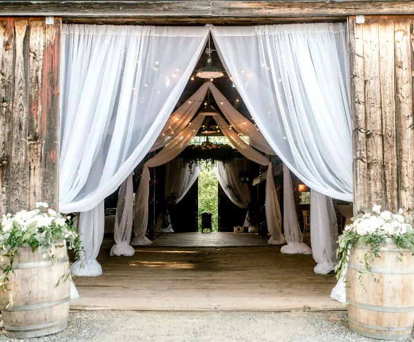 Interior of a rustic wooden barn venue decorated with white drapery, string lights, and floral arrangements.