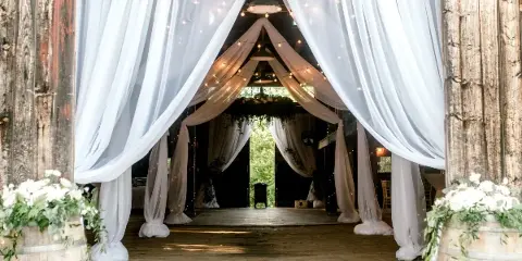 Interior of a rustic wooden barn venue decorated with white drapery, string lights, and floral arrangements.