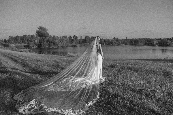 A black and white photograph of a bride in a long gown and veil standing in a grassy field near a body of water.
