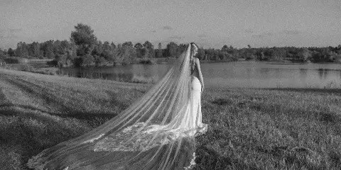 A black and white photograph of a bride in a long gown and veil standing in a grassy field near a body of water.