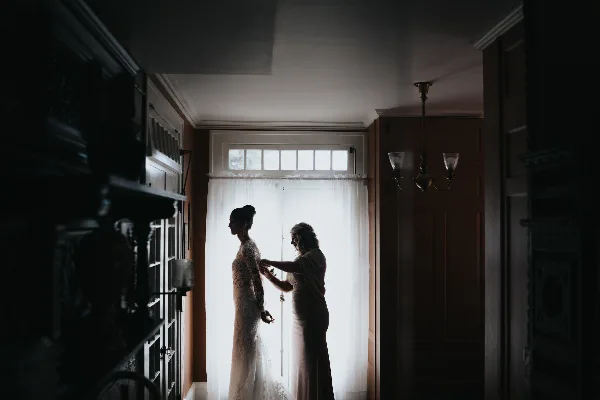 A bride in a lace gown stands in silhouette by a window while another woman adjusts her dress.