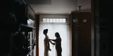 A bride in a lace gown stands in silhouette by a window while another woman adjusts her dress.