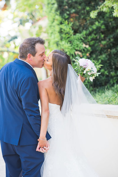 A bride and groom sharing a kiss outdoors, with the bride holding a bouquet of flowers.