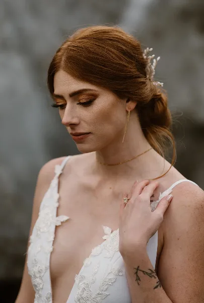 A bride with red hair styled in an updo with a floral accessory, wearing a white lace wedding dress and professional makeup.
