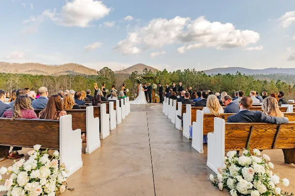 Outdoor wedding ceremony with a bride and groom at an altar, surrounded by guests in wooden pews and a mountain landscape in the background.