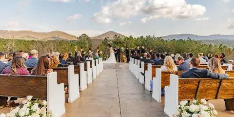 Outdoor wedding ceremony with a bride and groom at an altar, surrounded by guests in wooden pews and a mountain landscape in the background.