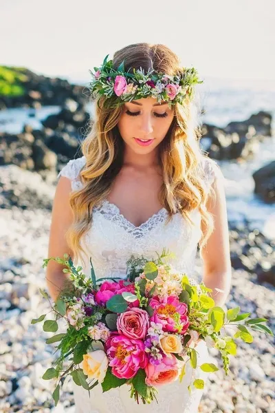 A bride with styled hair and makeup wearing a floral crown and holding a bouquet on a beach.