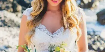 A bride with styled hair and makeup wearing a floral crown and holding a bouquet on a beach.