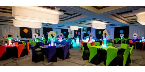 A large event hall set up with round tables, colorful chair covers, and decorative lighting.