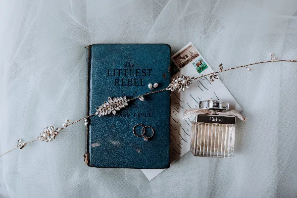 A flat lay photograph showing a vintage blue book titled 'The Littlest Rebel', a pair of wedding rings, a calligraphy invitation, and a decorative beaded hairpiece on a white tulle background.