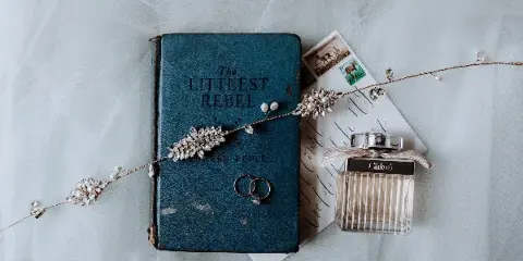 A flat lay photograph showing a vintage blue book titled 'The Littlest Rebel', a pair of wedding rings, a calligraphy invitation, and a decorative beaded hairpiece on a white tulle background.