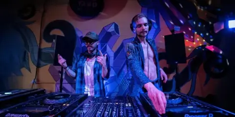 Two DJs performing at a live event with Pioneer DJ equipment and stage lighting.