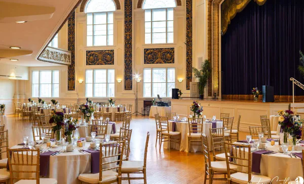 A formal event reception room with round tables set with white linens, purple runners, floral centerpieces, and gold chiavari chairs.