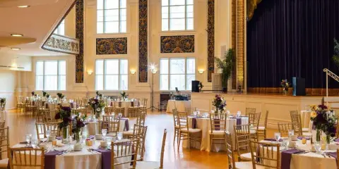 A formal event reception room with round tables set with white linens, purple runners, floral centerpieces, and gold chiavari chairs.
