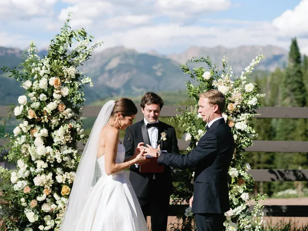 A bride and groom exchanging rings during an outdoor wedding ceremony set against a mountain landscape.