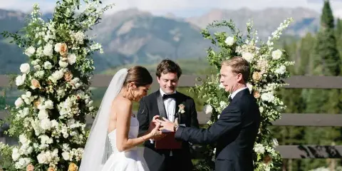 A bride and groom exchanging rings during an outdoor wedding ceremony set against a mountain landscape.