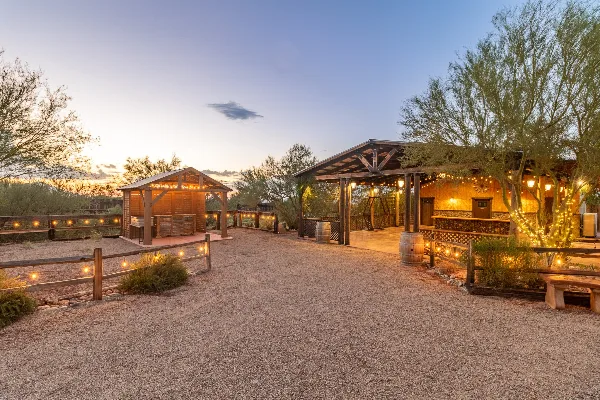 A rustic wooden event pavilion and gazebo illuminated by string lights at dusk in a desert setting.
