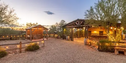 A rustic wooden event pavilion and gazebo illuminated by string lights at dusk in a desert setting.