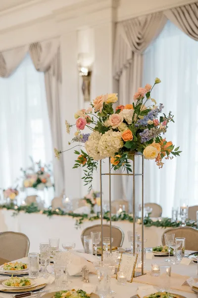 A tall floral centerpiece featuring pink, yellow, and white flowers on a gold stand, set on a decorated wedding table.
