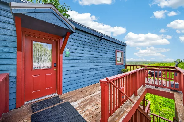 Exterior view of a blue cabin with a red door and deck overlooking a wide green landscape.